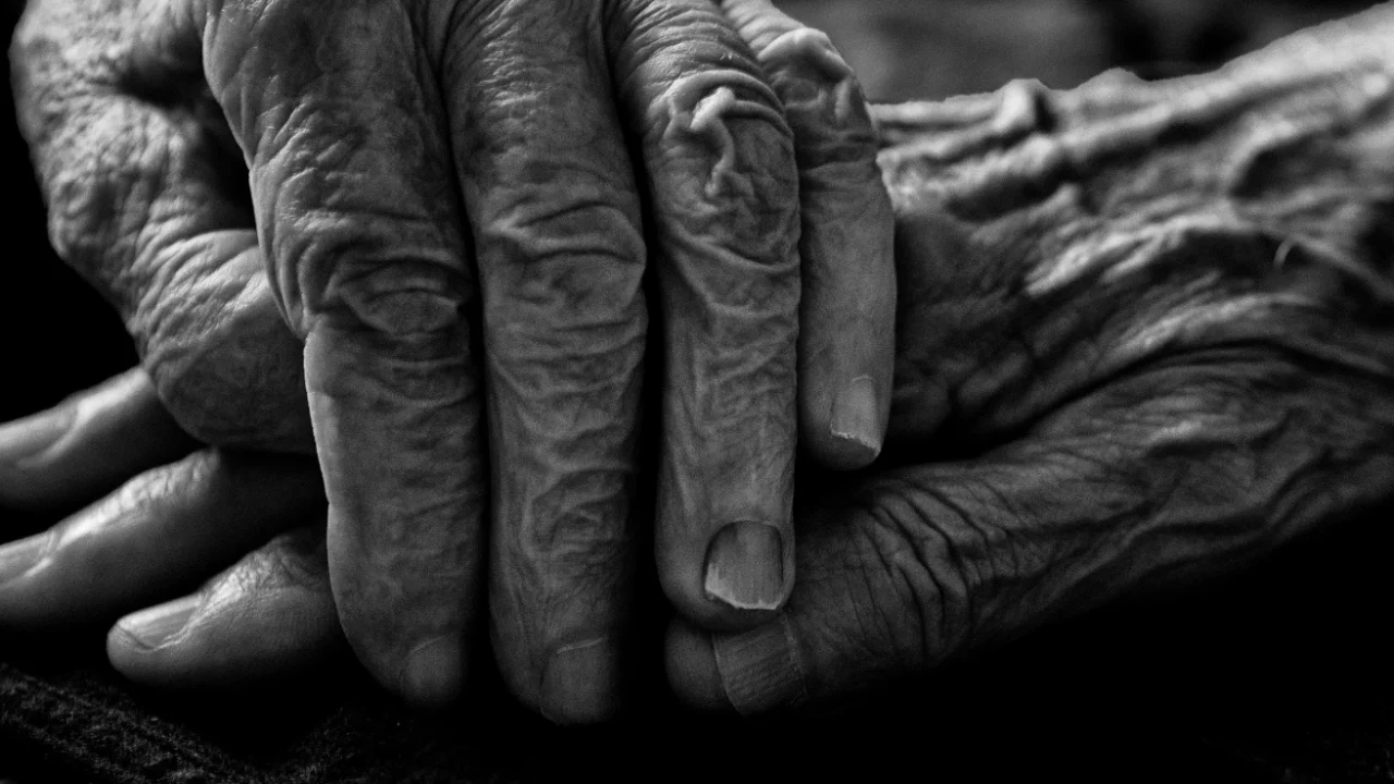 Close-up black-and-white image of an elderly person’s wrinkled hands resting together in their lap.