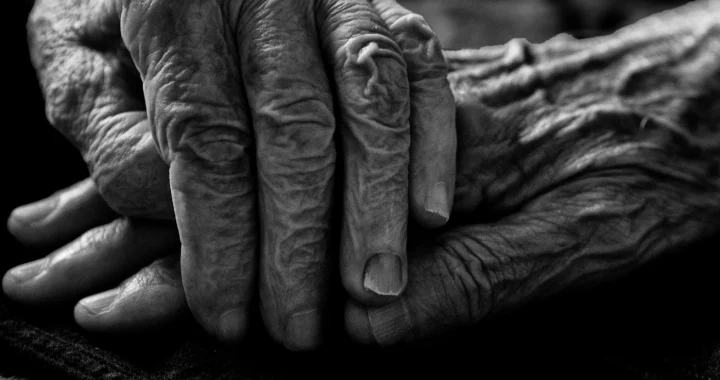 Close-up black-and-white image of an elderly person’s wrinkled hands resting together in their lap.