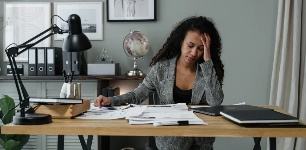 A woman sits at a desk covered with papers and documents, holding her head in one hand while looking stressed. A laptop, desk lamp, and office items surround her in a workspace.
