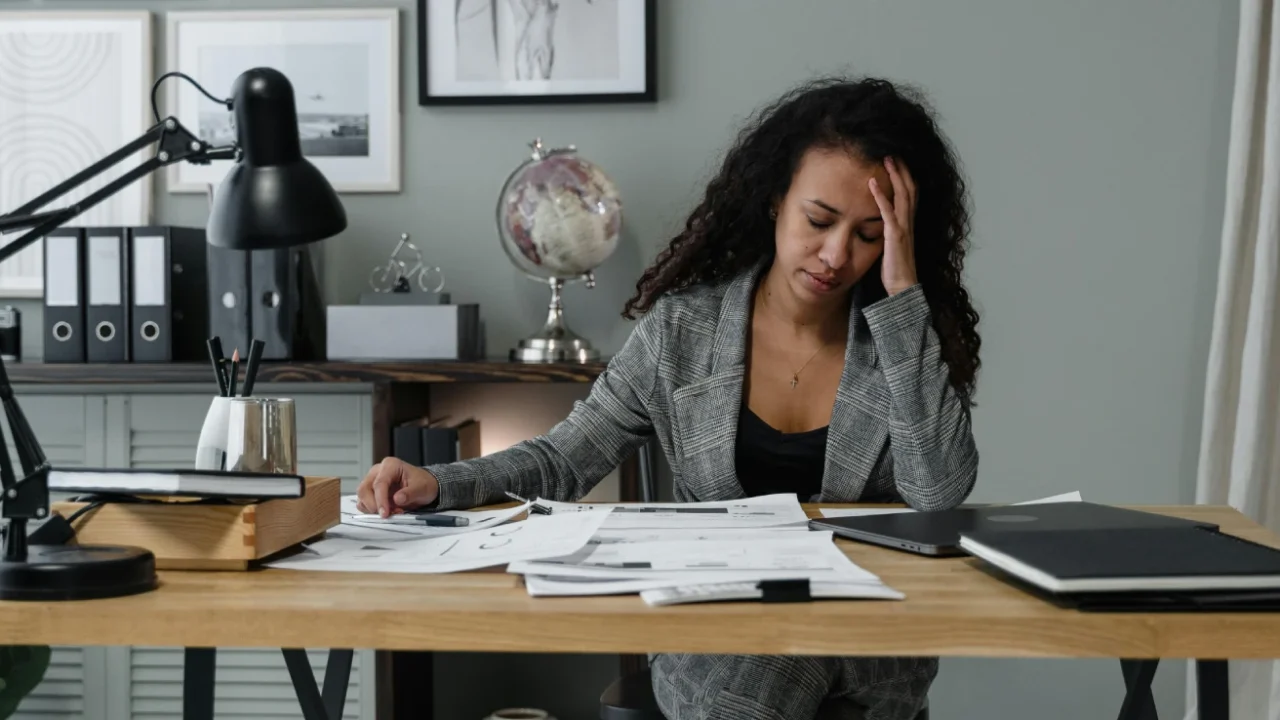 A woman sits at a desk covered with papers and documents, holding her head in one hand while looking stressed. A laptop, desk lamp, and office items surround her in a workspace.
