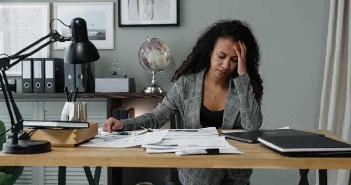 A woman sits at a desk covered with papers and documents, holding her head in one hand while looking stressed. A laptop, desk lamp, and office items surround her in a workspace.
