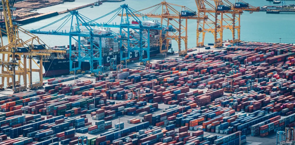 Aerial view of a busy cargo port with rows of stacked shipping containers, towering cranes, and container ships docked beside the harbor.