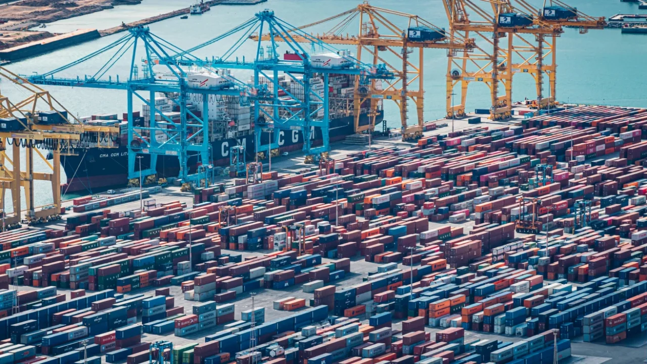 Aerial view of a busy cargo port with rows of stacked shipping containers, towering cranes, and container ships docked beside the harbor.