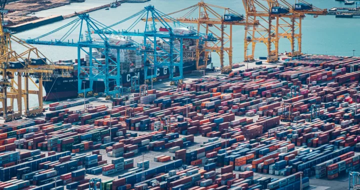 Aerial view of a busy cargo port with rows of stacked shipping containers, towering cranes, and container ships docked beside the harbor.