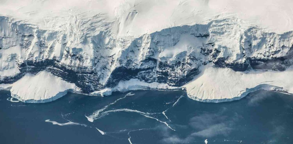 Aerial view of an Antarctic coastline, with steep snow-covered cliffs dropping to a dark blue ocean dotted with floating sea ice and thin cracks in the surface.