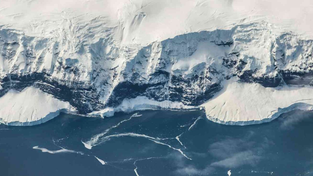 Aerial view of an Antarctic coastline, with steep snow-covered cliffs dropping to a dark blue ocean dotted with floating sea ice and thin cracks in the surface.