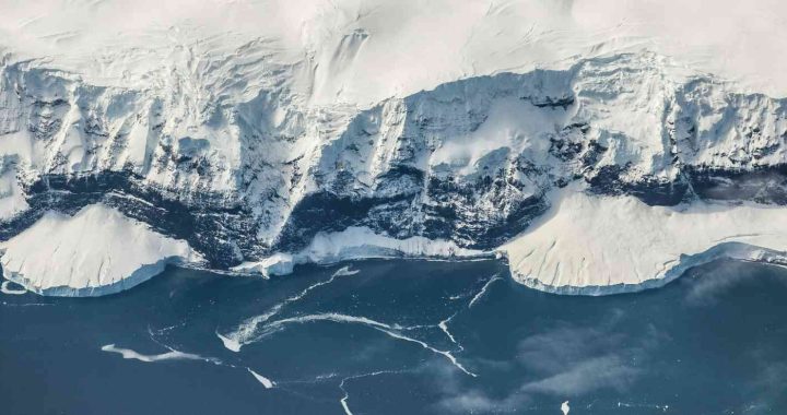 Aerial view of an Antarctic coastline, with steep snow-covered cliffs dropping to a dark blue ocean dotted with floating sea ice and thin cracks in the surface.