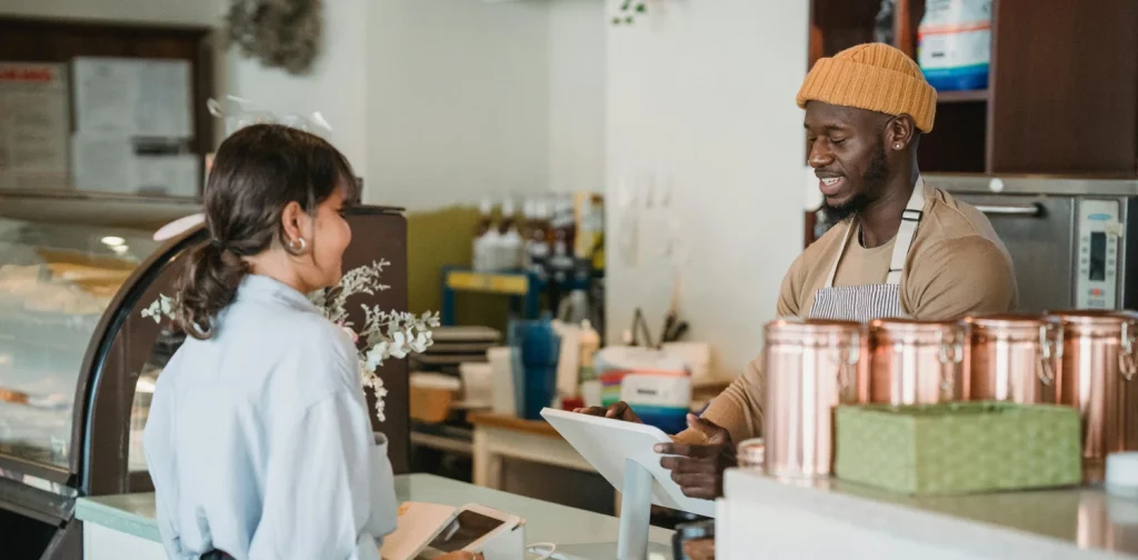 A cafe worker wearing a mustard beanie and apron smiles while taking an order from a customer at the counter. The customer stands beside a card payment machine, and copper containers and a pastry display case are visible in the cozy cafe interior.