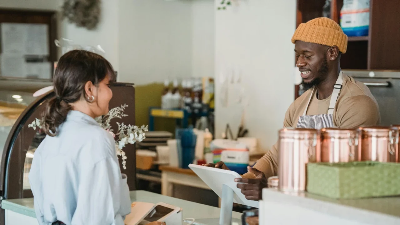 A cafe worker wearing a mustard beanie and apron smiles while taking an order from a customer at the counter. The customer stands beside a card payment machine, and copper containers and a pastry display case are visible in the cozy cafe interior.