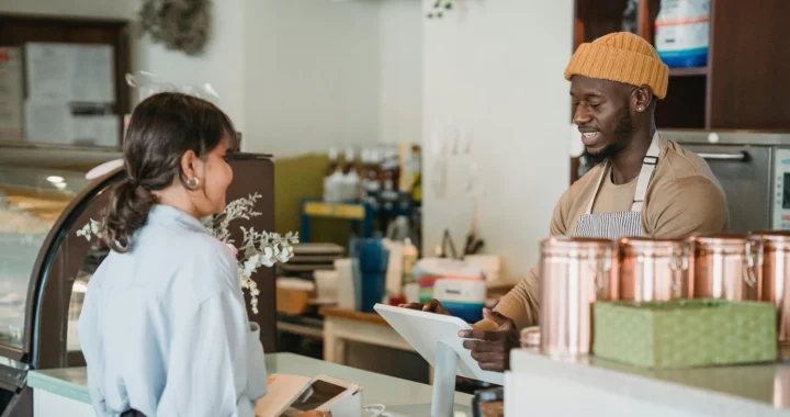 A cafe worker wearing a mustard beanie and apron smiles while taking an order from a customer at the counter. The customer stands beside a card payment machine, and copper containers and a pastry display case are visible in the cozy cafe interior.
