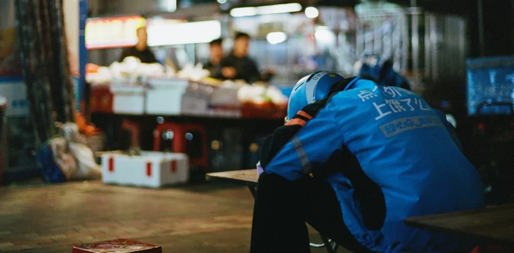 A delivery rider wearing a blue Ele.me jacket and helmet rests his head on a counter at a night market, appearing worn out mid-shift.