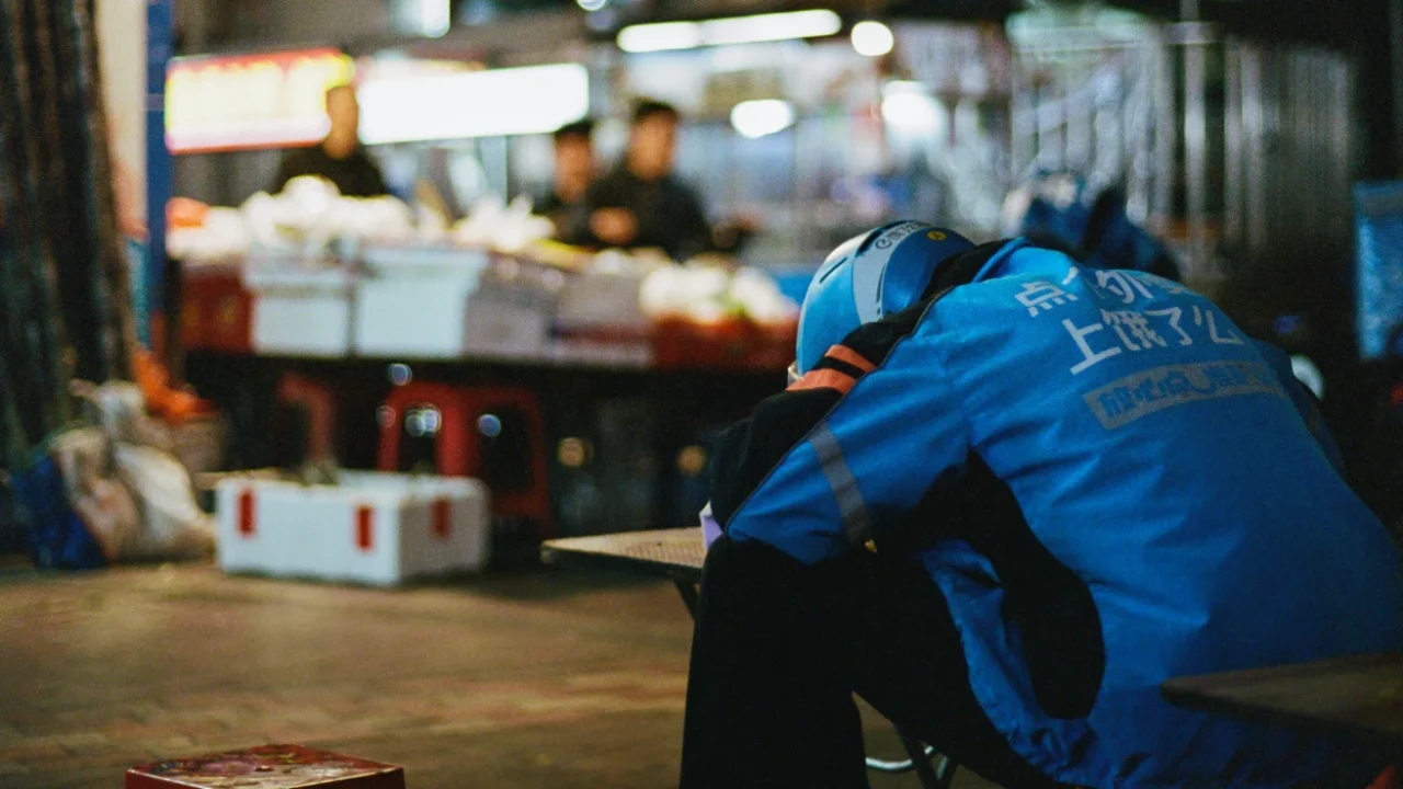 A delivery rider wearing a blue Ele.me jacket and helmet rests his head on a counter at a night market, appearing worn out mid-shift.