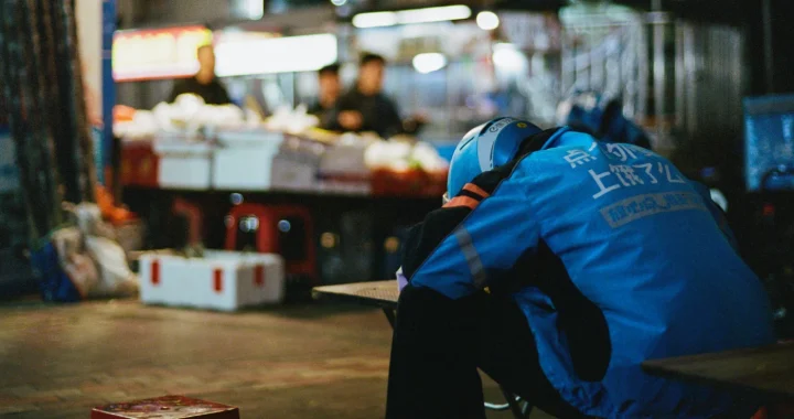 A delivery rider wearing a blue Ele.me jacket and helmet rests his head on a counter at a night market, appearing worn out mid-shift.