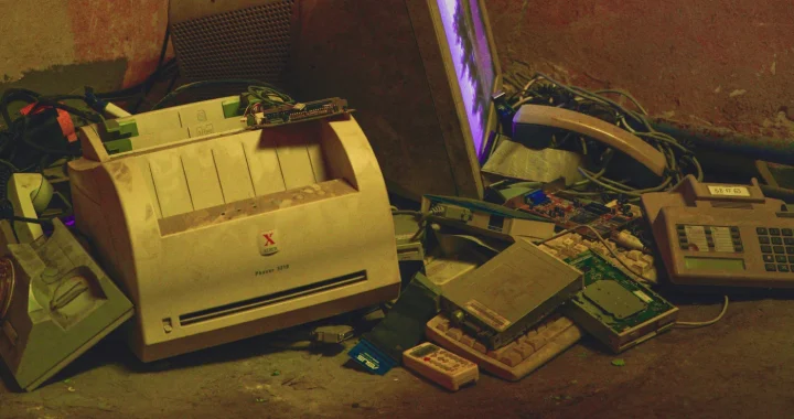 Discarded electronic devices, including a CRT monitor, printer, keyboard, and telephone, piled together as electronic waste.