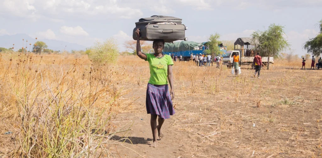 A woman carries a large suitcase on her head while walking across a dry rural landscape. In the background, people gather near trucks and vehicles.