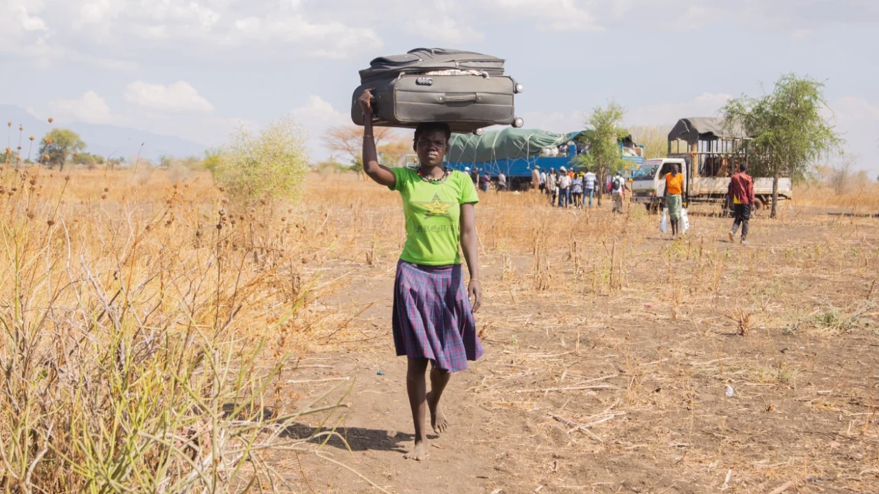 A woman carries a large suitcase on her head while walking across a dry rural landscape. In the background, people gather near trucks and vehicles.