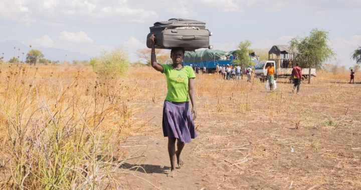 A woman carries a large suitcase on her head while walking across a dry rural landscape. In the background, people gather near trucks and vehicles.