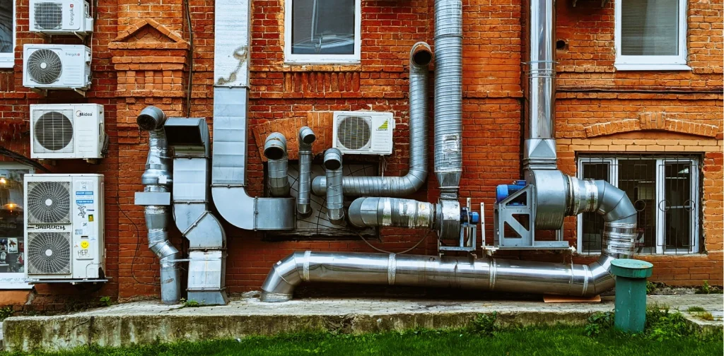 Exterior wall of a red brick building with multiple wall-mounted air conditioning units, large silver ventilation and exhaust pipes, and several windows.