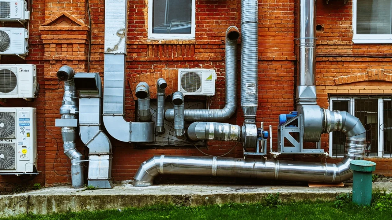 Exterior wall of a red brick building with multiple wall-mounted air conditioning units, large silver ventilation and exhaust pipes, and several windows.