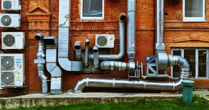 Exterior wall of a red brick building with multiple wall-mounted air conditioning units, large silver ventilation and exhaust pipes, and several windows.