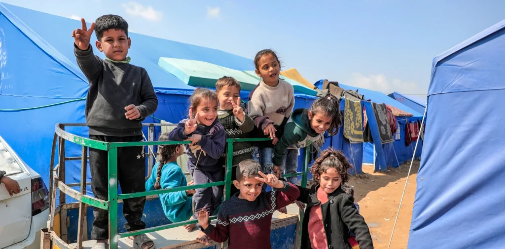 A group of children smiling, waving, and making peace signs while standing on a small trailer in a camp with rows of blue tents under a clear sky.