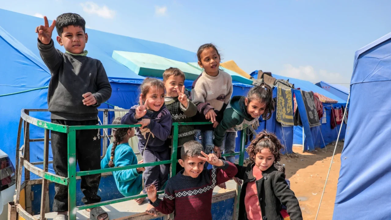 A group of children smiling, waving, and making peace signs while standing on a small trailer in a camp with rows of blue tents under a clear sky.
