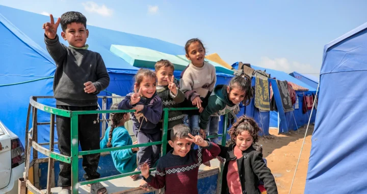 A group of children smiling, waving, and making peace signs while standing on a small trailer in a camp with rows of blue tents under a clear sky.