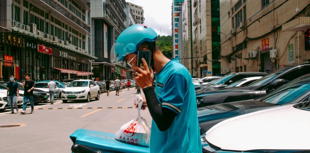 a delivery worker in blue helmet and blue jacket standing beside cars while talking on the phone