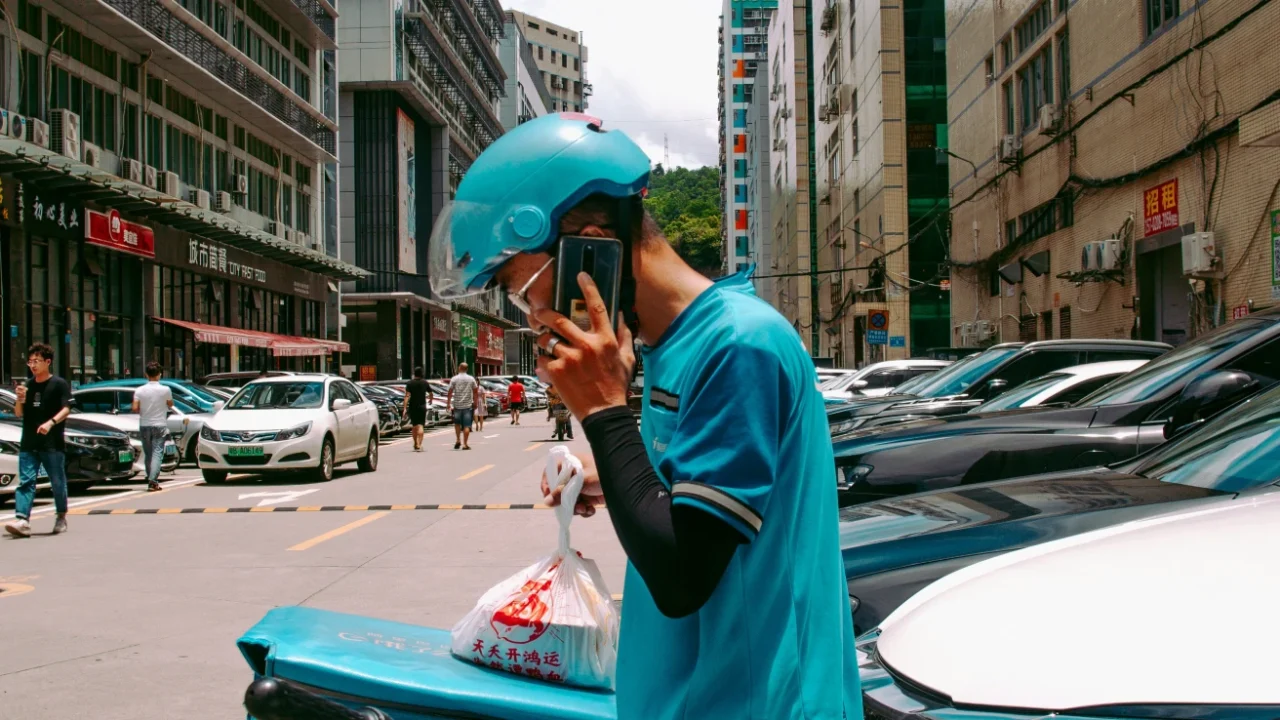 a delivery worker in blue helmet and blue jacket standing beside cars while talking on the phone