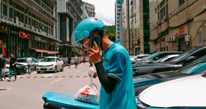 a delivery worker in blue helmet and blue jacket standing beside cars while talking on the phone