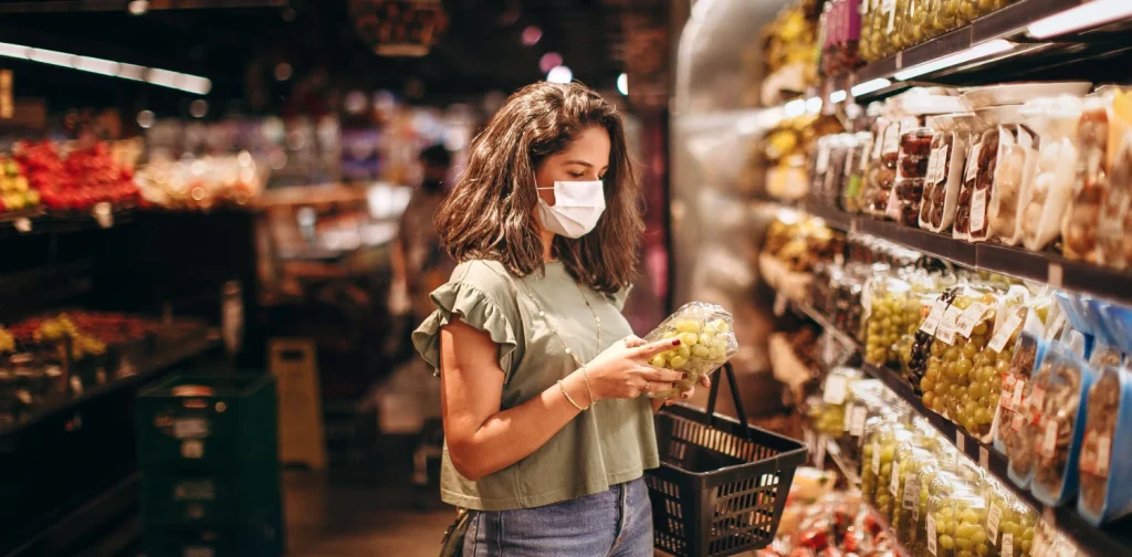 Young woman in a mask selecting fresh fruits in a supermarket produce aisle.