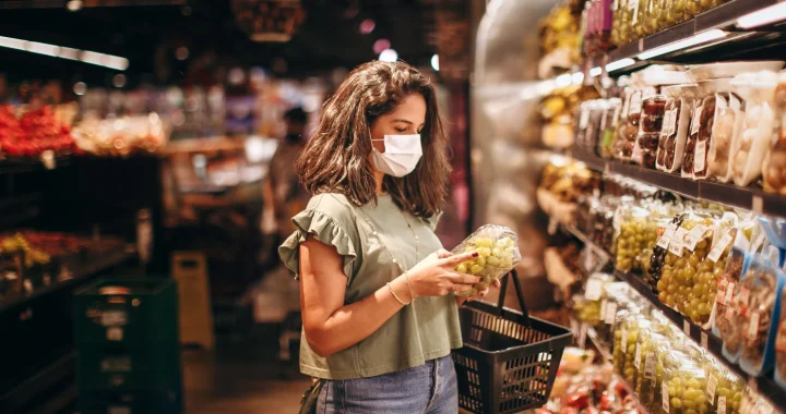 Young woman in a mask selecting fresh fruits in a supermarket produce aisle.