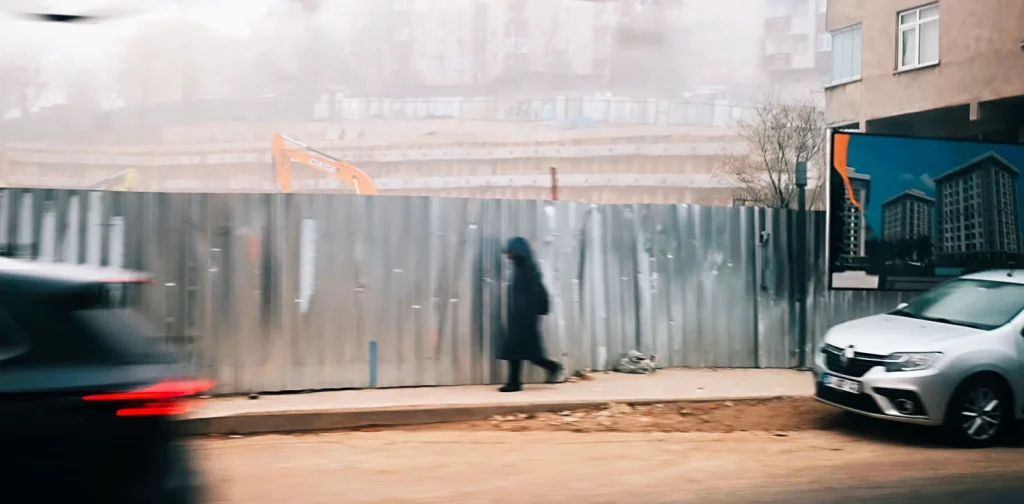 a pedestrian walking at the sidewalk with construction by the side of the road and car traffic on the street