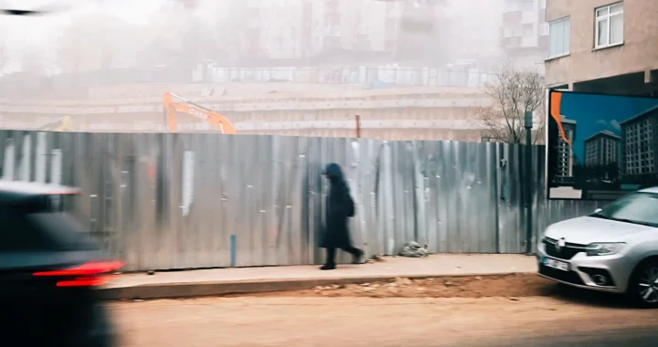 a pedestrian walking at the sidewalk with construction by the side of the road and car traffic on the street