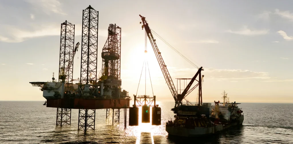 Offshore oil drilling platform and construction vessel operating at sea during sunset with a large care lifting equipment.