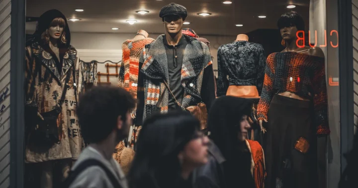 A clothing store displays trendy outfits on mannequins with people passing by the store.